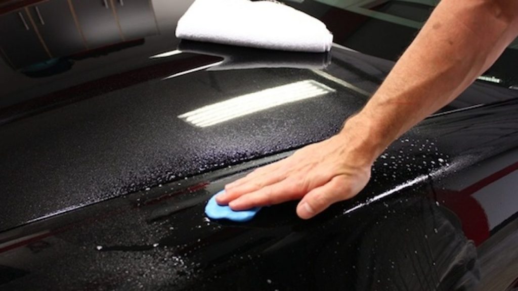 Main utilisant un tampon de polissage bleu sur le toit d'une voiture pour appliquer un produit de décontamination de la peinture.