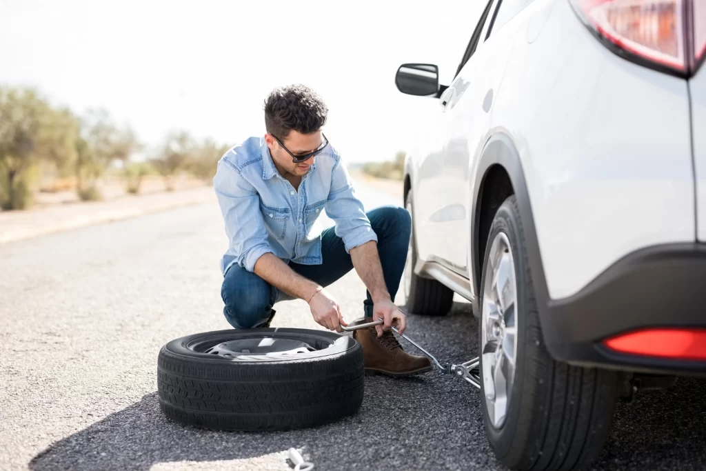 Homme accroupi en train de réparer un pneu de voiture avec un cric à proximité sous un soleil éclatant.