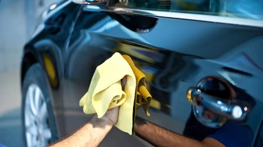 Main tenant un chiffon en microfibre jaune lustrant la carrosserie bleue métallisée d'une voiture pour un nettoyage minutieux.